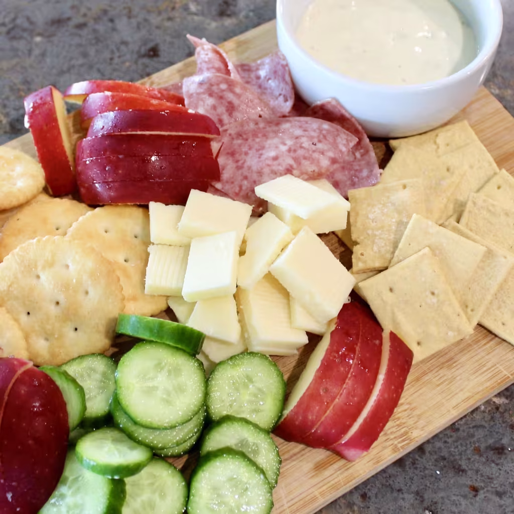 Board with crackers, cheese, veggies, meat, apples, and dipping sauce